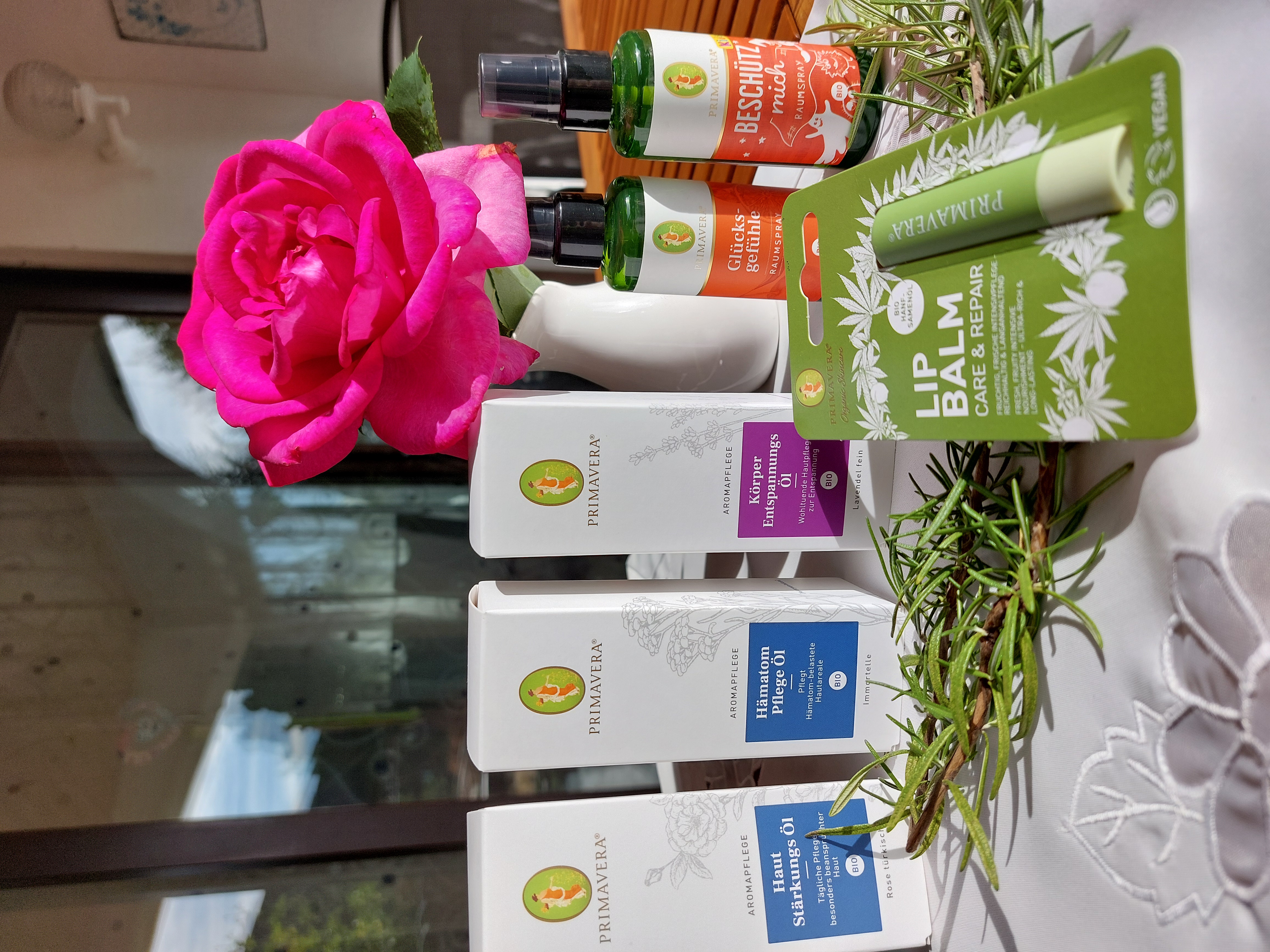 eine kleine Rose in einer Vase, dahinter drei Schachteln und 2 Fläschchen von Primavera. Im Vordergrund ein Lippenstift mit der Aufschrift "Lip Balm"